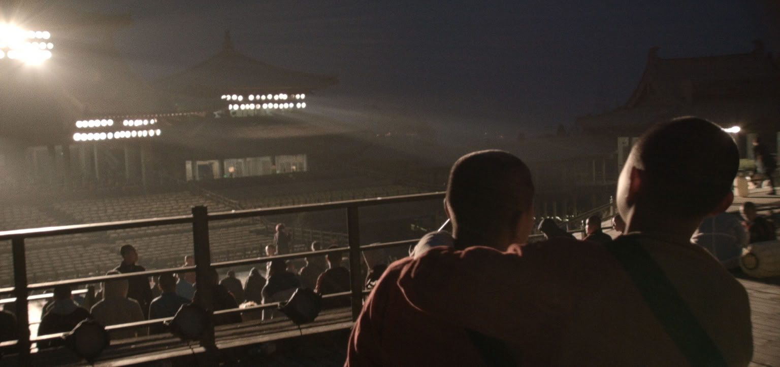 Two young boys in robes stand on a stage for a kungfu show, facing a lit-up traditional building with a group of people sitting on the ground in the background.