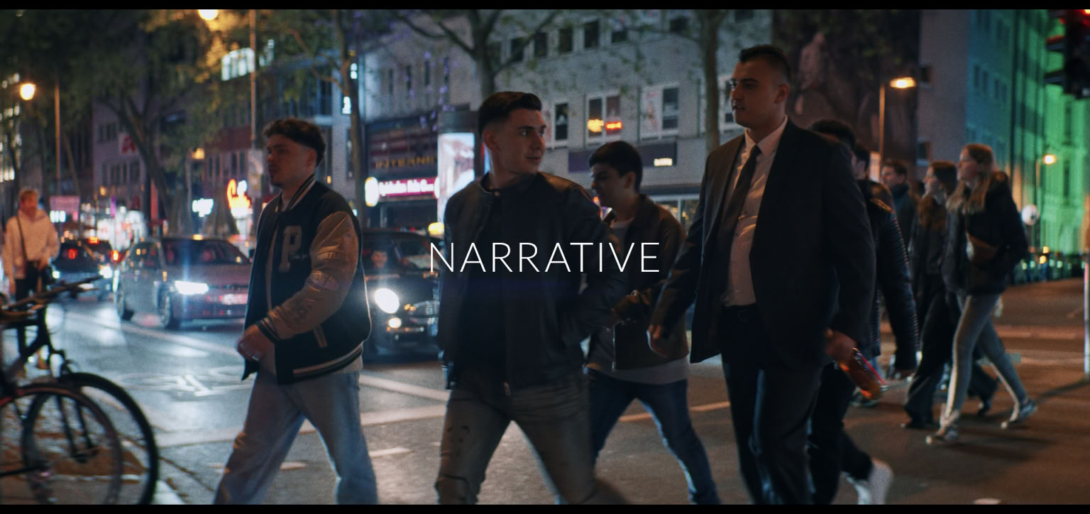 A group of people walking on a city street at night, with cars and bicycles nearby, and buildings illuminated in the background.