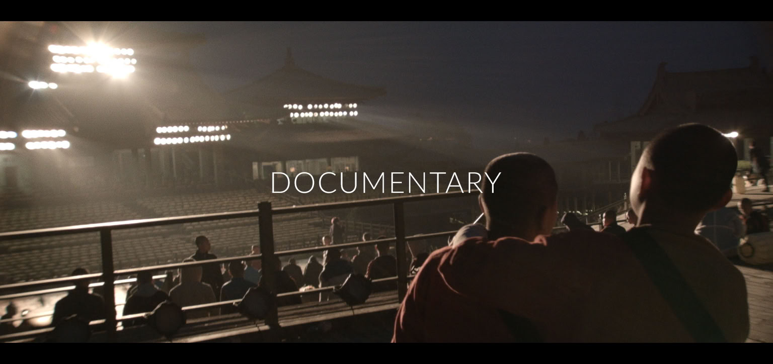 Two young boys in robes stand on a stage for a kungfu show, facing a lit-up traditional building with a group of people sitting on the ground in the background.